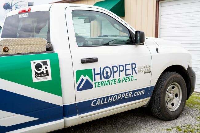 A white pickup truck branded with Hopper Termite & Pest, Inc. is parked next to a building. The truck displays a website CALLHOPPER.com and a QualityPro logo on the side.