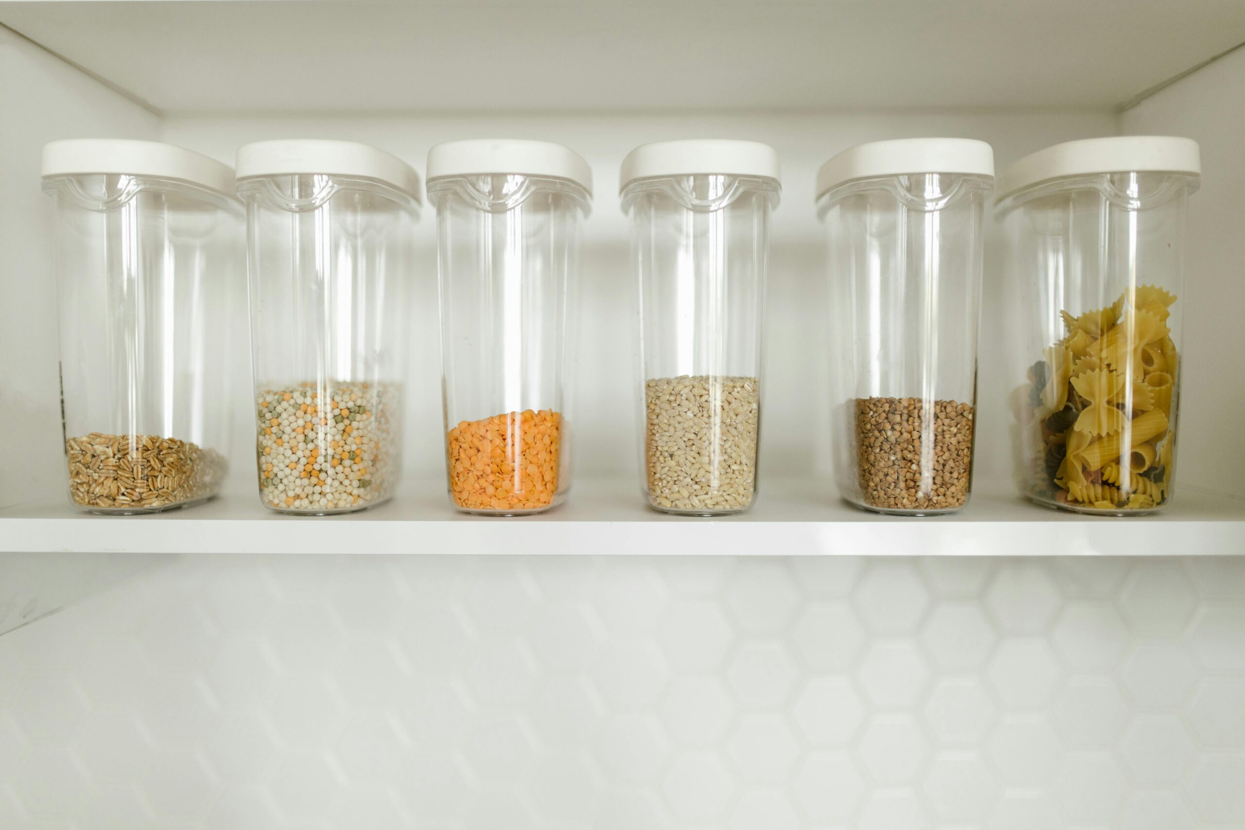 Six clear containers with white lids are neatly arranged on a white shelf, securely storing grains, lentils, and pasta safely away from spring pests. The pristine background features a stylish white hexagonal tile pattern.