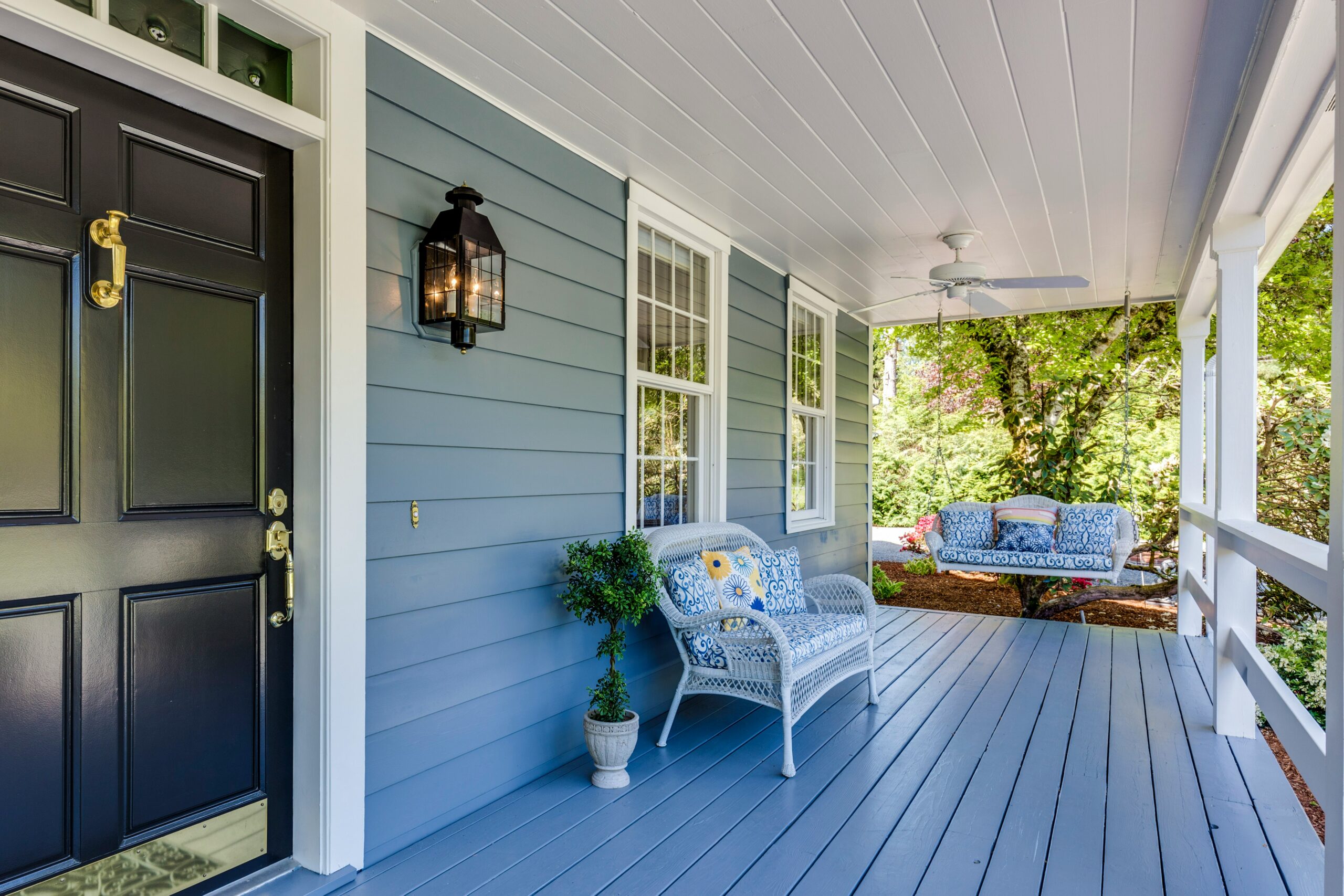 A cozy front porch with blue siding, a black door, wicker chairs with blue cushions, a potted plant, a ceiling fan, and greenery visible in the background—perfect for relaxing after tackling spring pest control.