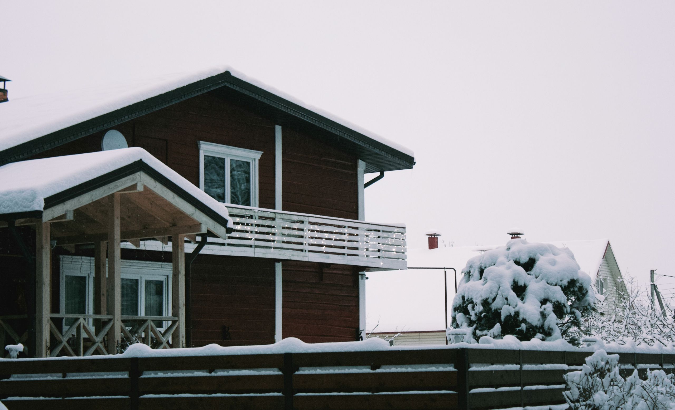pest barriers A wooden house with a balcony and porch is covered in snow. Snow blankets the fence, rooftop, pest barriers, trees, and surrounding area, creating a wintry scene under an overcast sky.