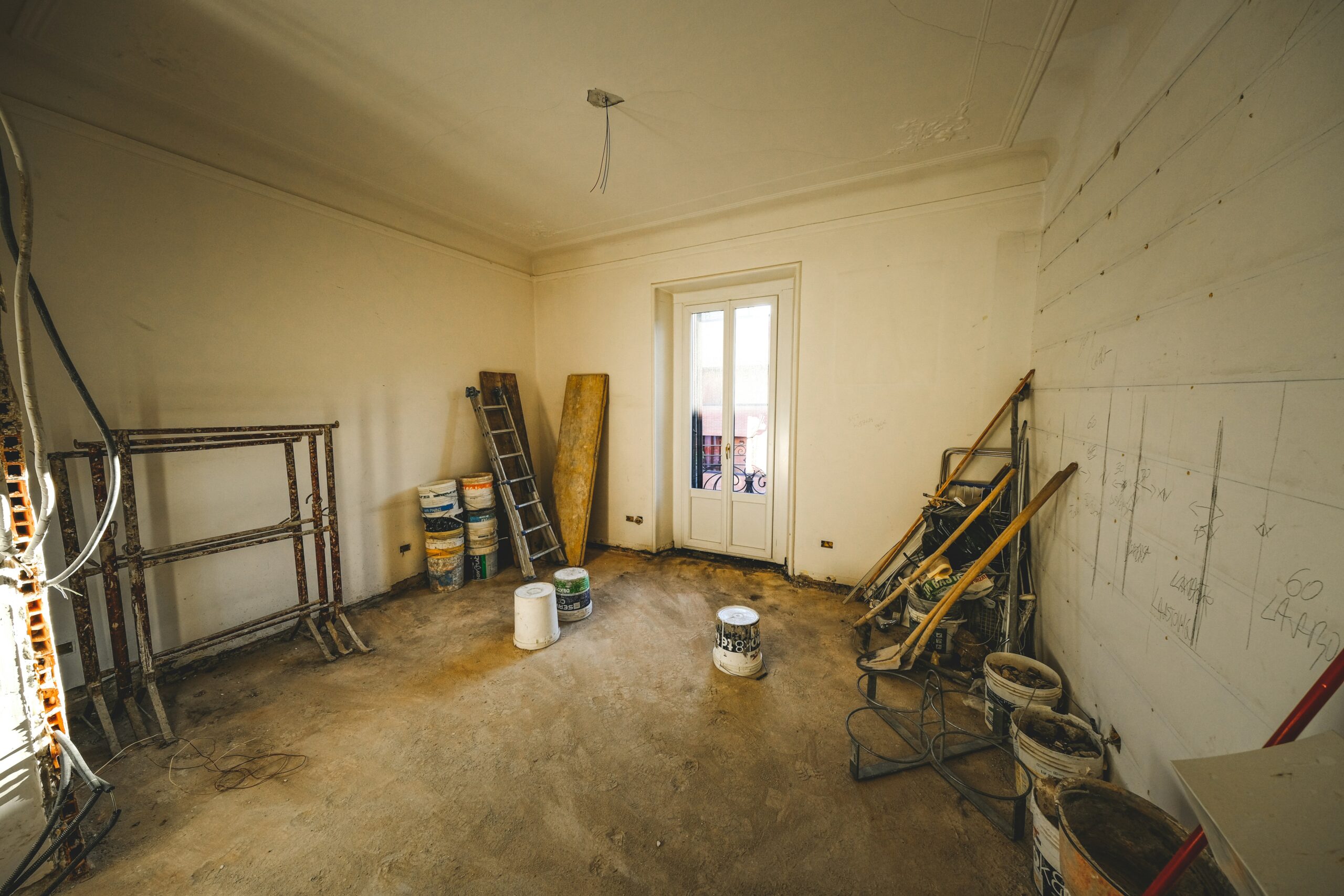 A room under renovation with bare walls, construction materials, paint cans, ladders, and scaffolding scattered around. Sunlight streams in through a double door leading to a balcony. The unfinished floor is dusty, ready for inspection for possible termite infestations.