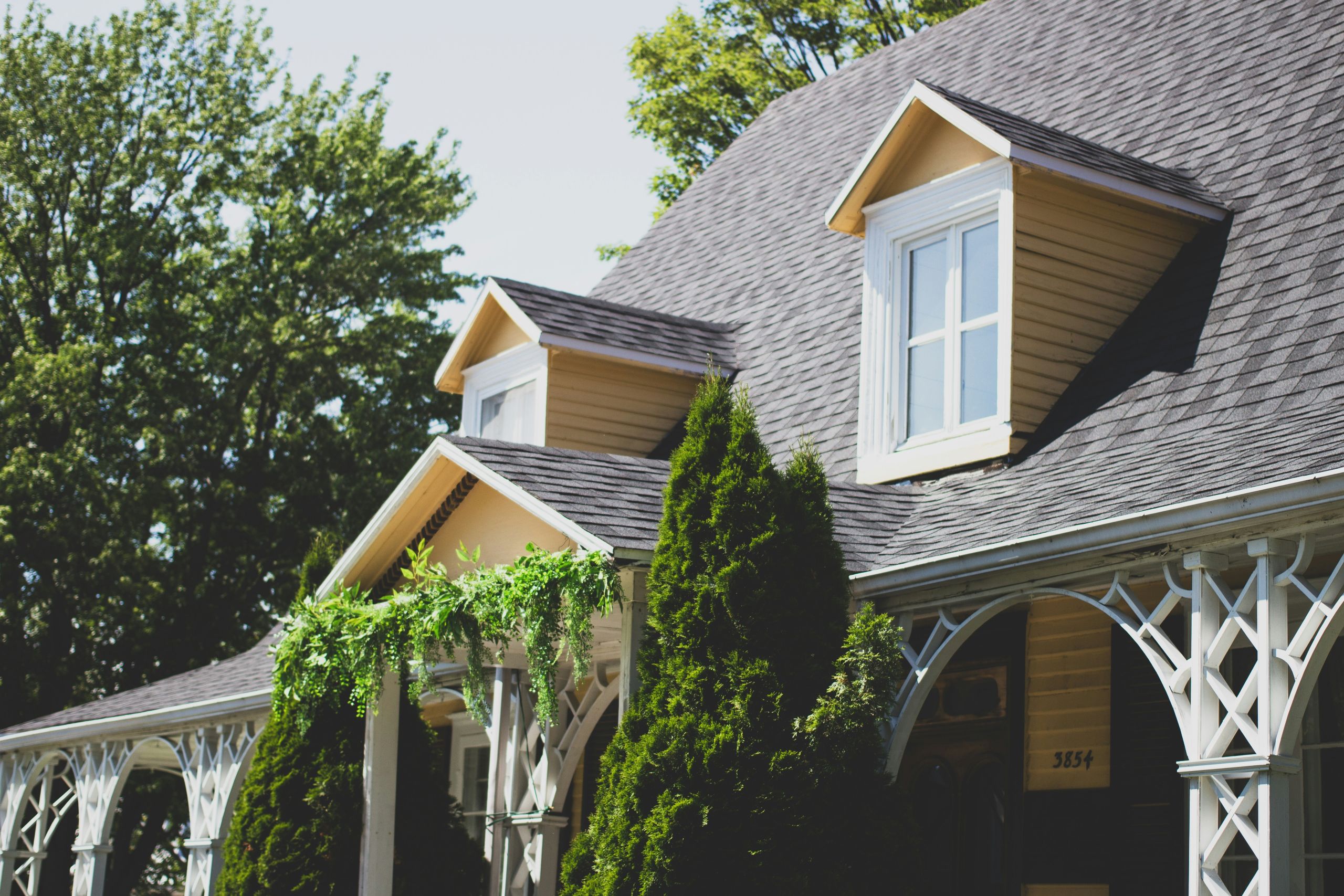 A yellow house with dormer windows, a dark shingled roof, and white lattice columns is surrounded by green trees and bushes—perfect for enjoying sunny days while keeping an eye out for common spring pests.