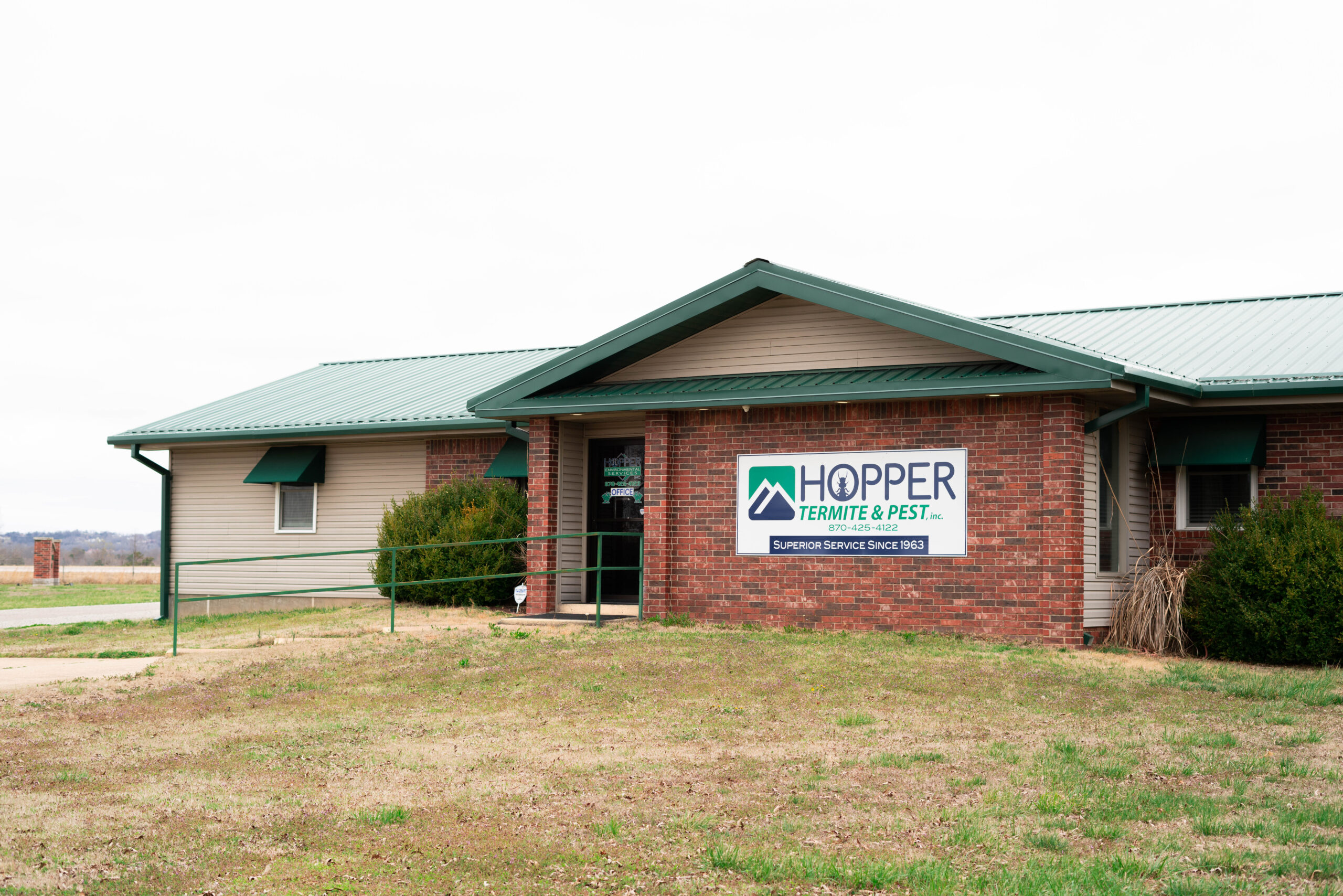 A single-story brick and siding building with a green roof and a large sign reading Hopper Termite & Pest on the front. The entrance has a ramp, and the building is surrounded by grass—ready to help with Common Spring Pest Problems.