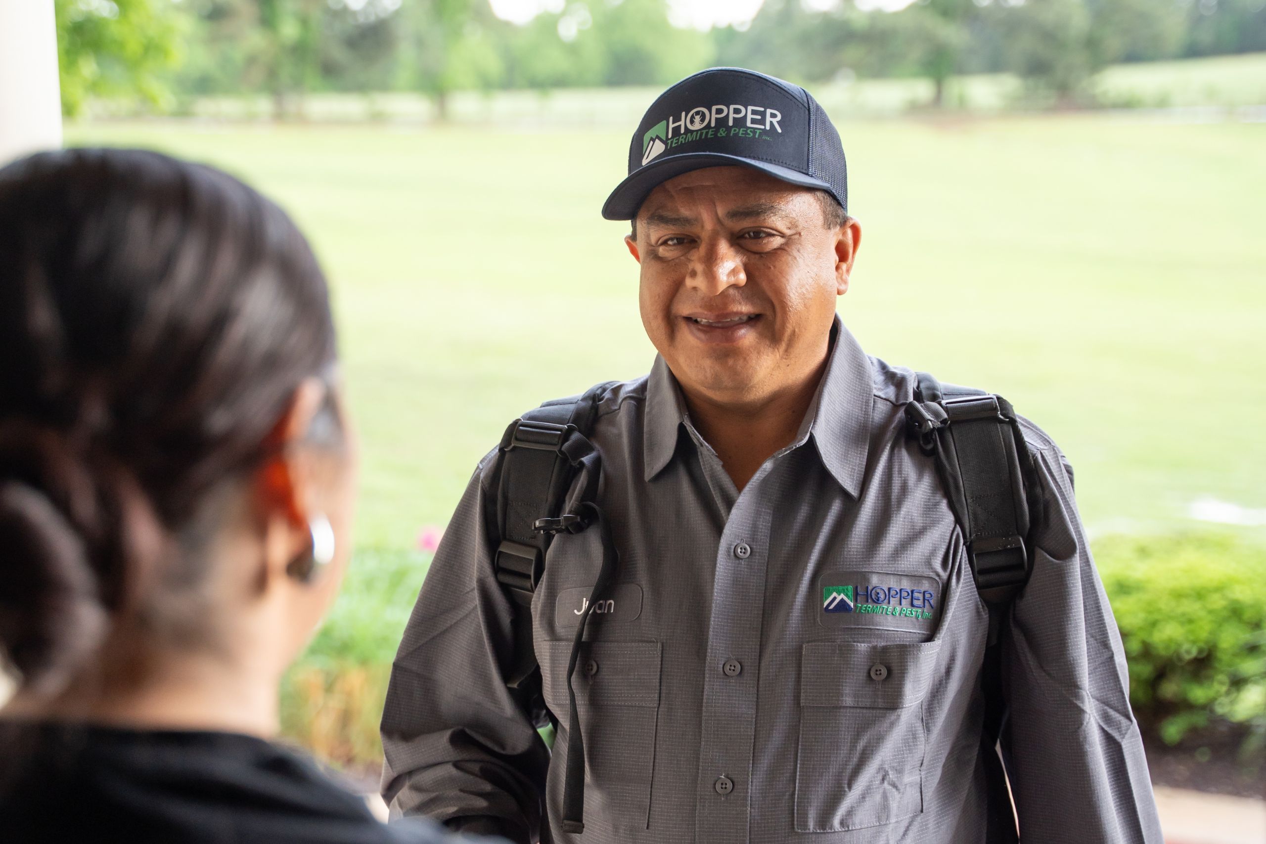 A smiling technician in a gray uniform and black cap with Hopper logo stands outside, carrying a backpack. He faces a woman whose back is to the camera, with greenery visible in the background.