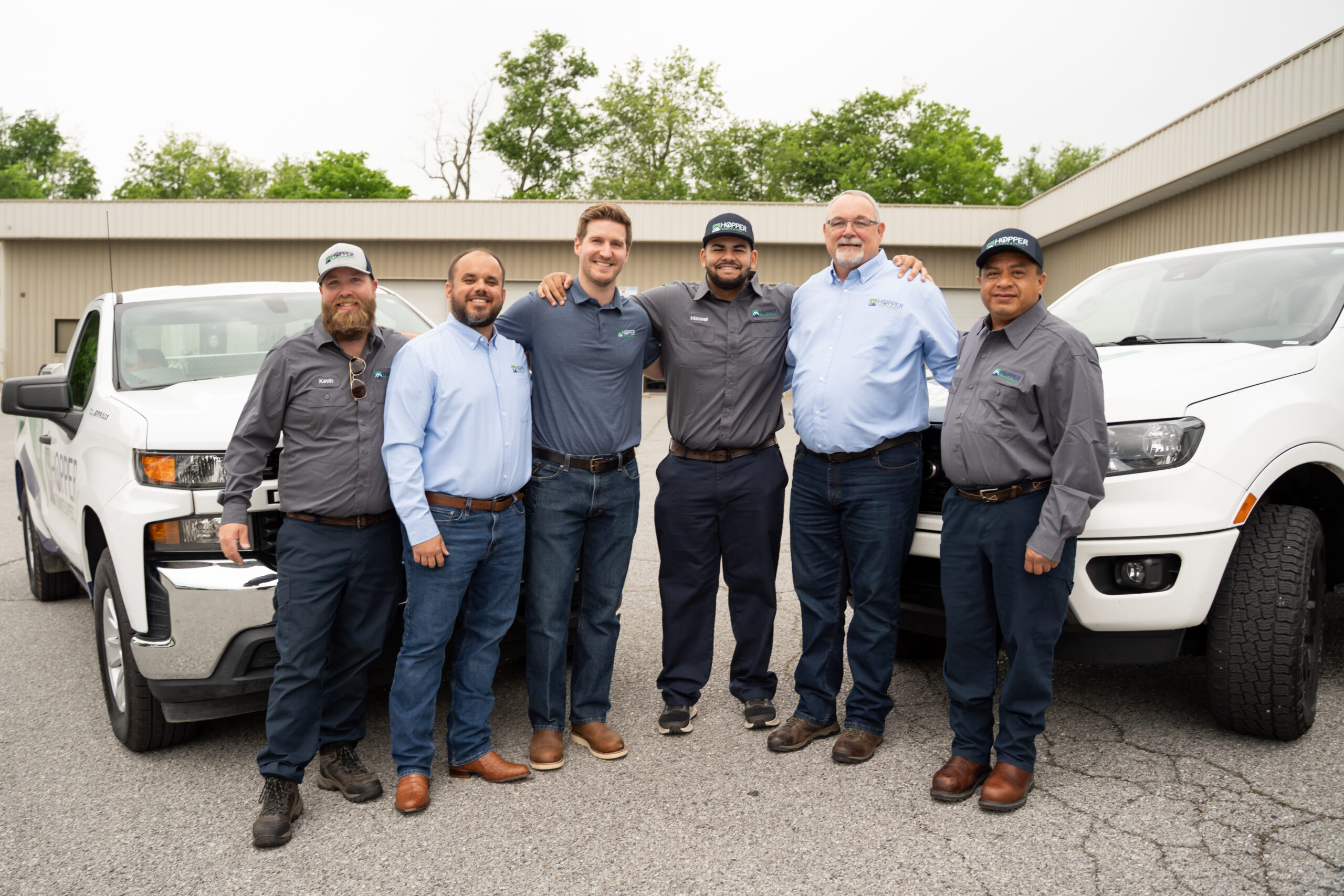 Six men wearing work shirts and jeans stand smiling between two white pickup trucks in a parking lot, with buildings and trees in the background.