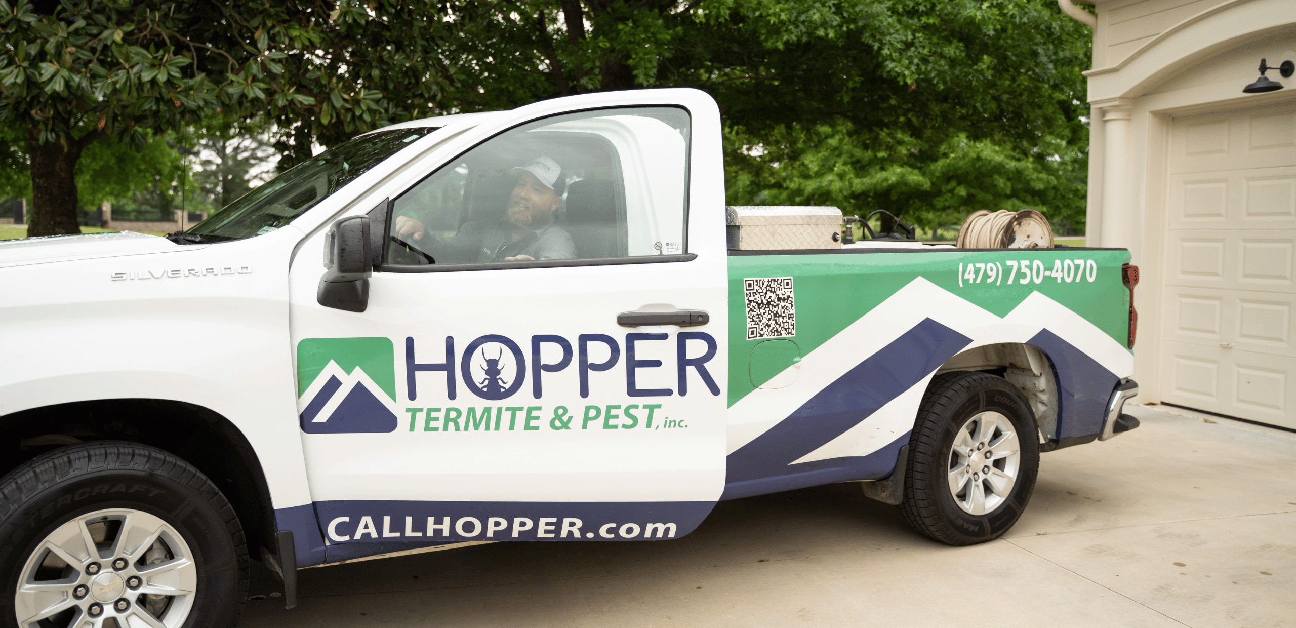A white pickup truck with Hopper Termite & Pest, Inc. branding, contact information, QR code, and logo is parked in a driveway; a man sits in the drivers seat.