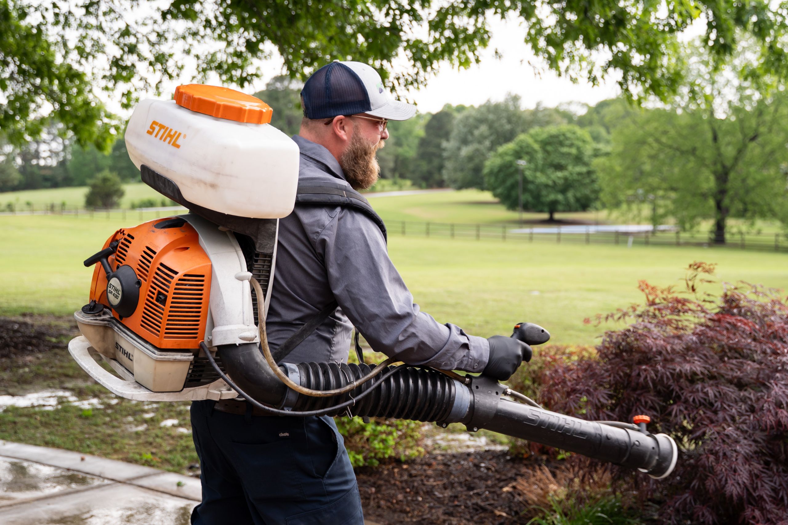 A man wearing a cap and gloves uses a STIHL leaf blower in a park-like setting with green grass, trees, and a cloudy sky in the background.