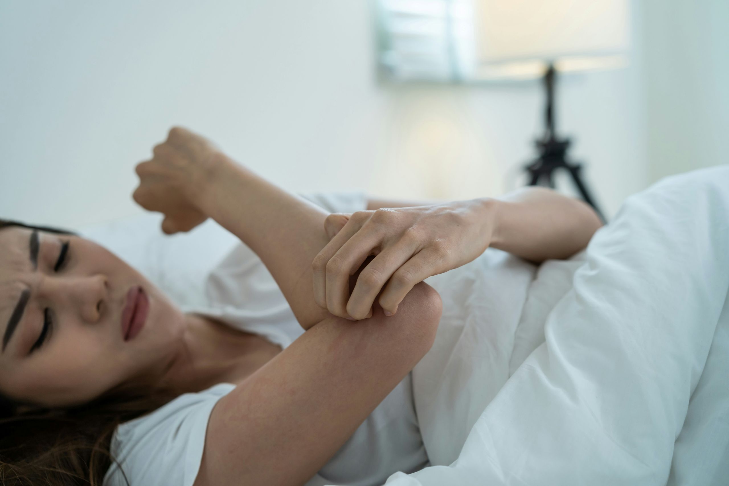fleas, ticks, and mosquitoes A young woman lies in bed, scratching her arm with an expression of discomfort.