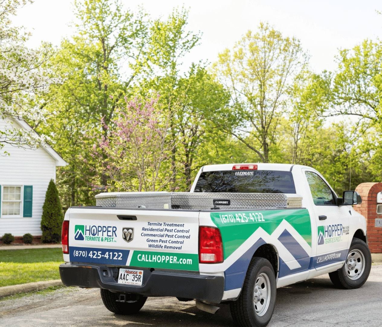 A white and green Hopper Pest Control truck is parked in a residential driveway, with trees and a white house in the background. The truck displays contact info and pest control services.