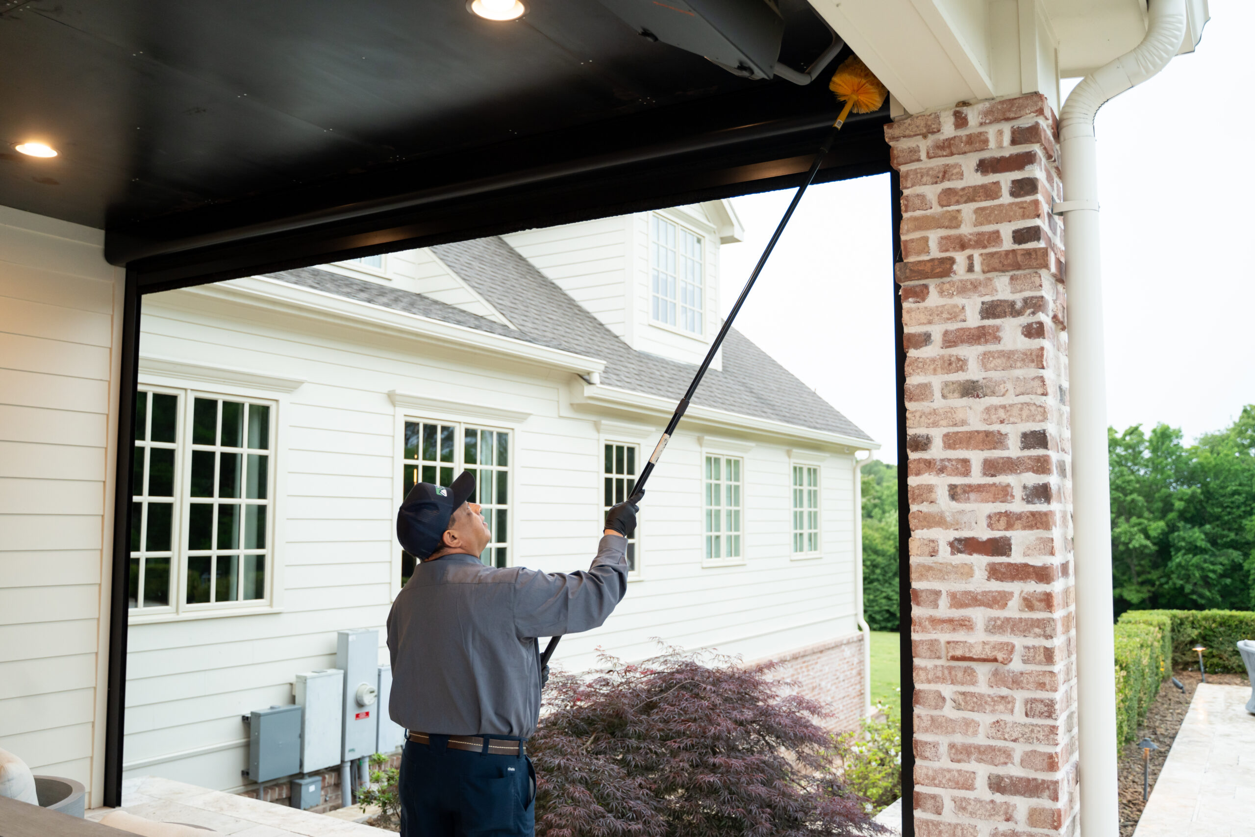 Preparing Your Home for Fall Pest Activity A person uses a long-handled duster to clean the upper frame of a patio door, standing outside a house with light-colored siding and large windows—a routine that can help prevent fall pest activity and reduce the need for ant pest control services.