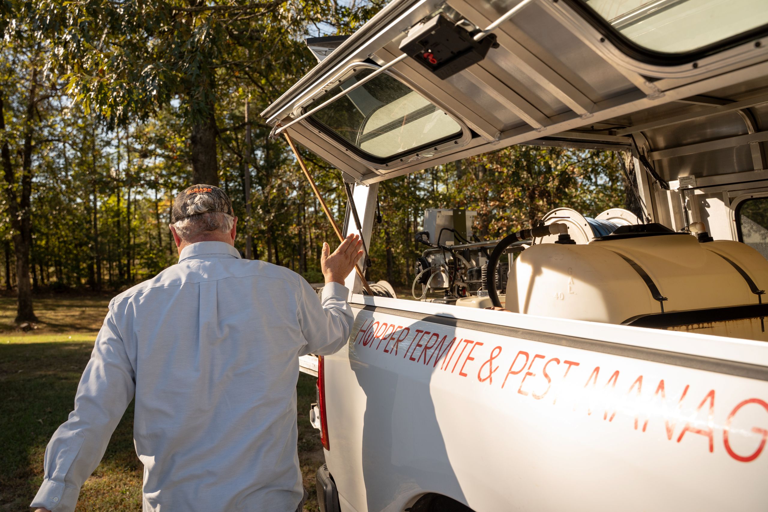 A man in a cap and light shirt walks toward a white pest control truck with equipment in the back, parked in a sunlit wooded area.